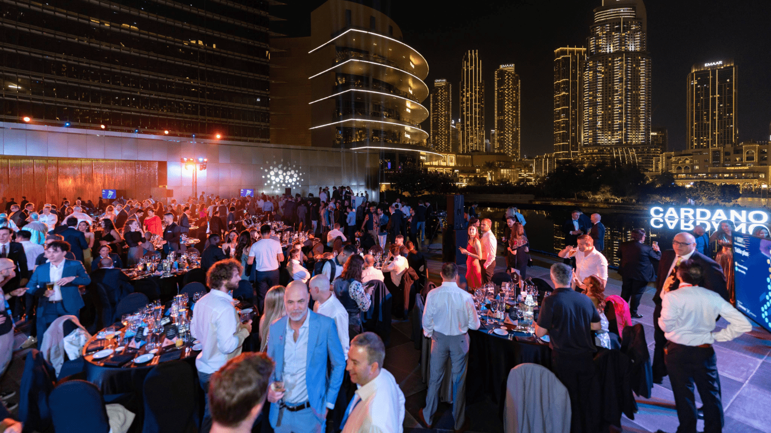 A large group of people in formal attire attend the Cardano Summit 2023 outdoor evening event with round dinner tables, city skyscrapers, and illuminated buildings in the background. Some guests mingle or sit at their tables.