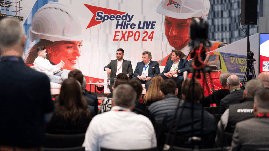 A panel of four men in business attire speak on stage at Speedy Hire Live Expo, with an audience watching and a large banner featuring construction workers in the background.