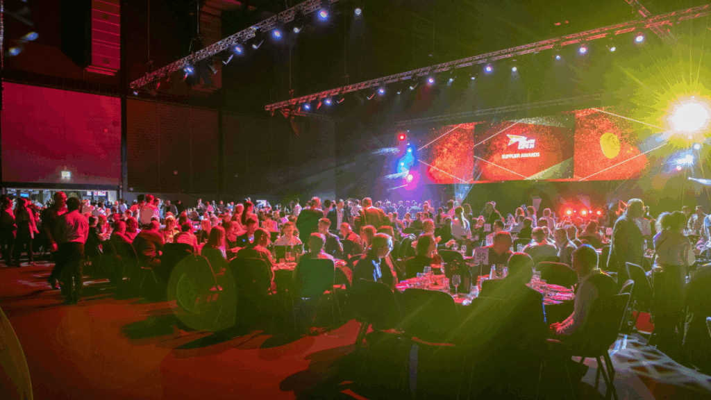 A large crowd in formal attire sits at round tables in a dimly lit event hall, as bright stage lights illuminate the “SUPPLY AWARDS” screen during the Speedy Hire Live Expo.