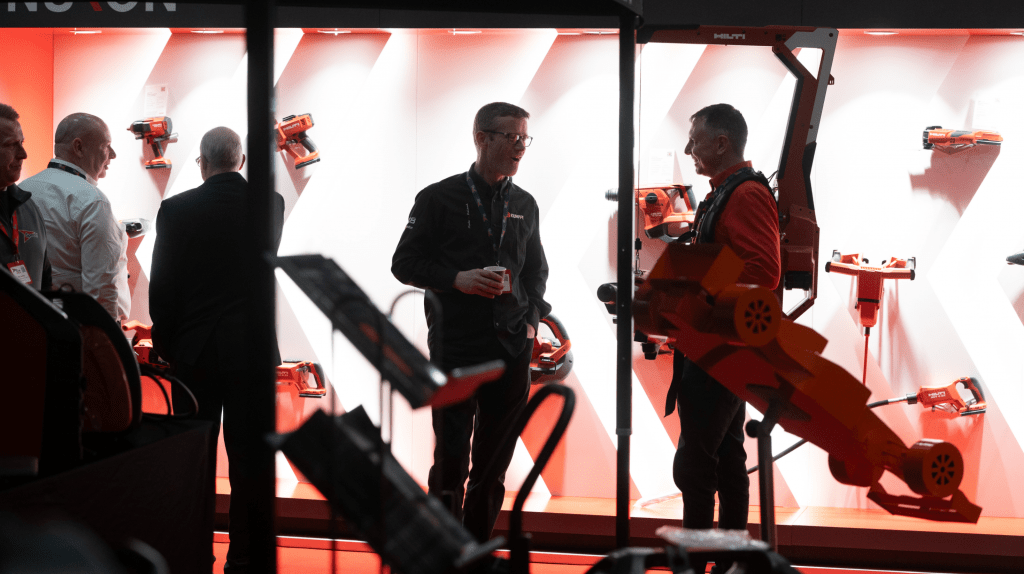 Two men converse in front of a brightly lit display of orange power tools at a Live Expo exhibit. Three other men in business attire stand nearby, with mounted tools showcased on a striking white and red wall in the background.