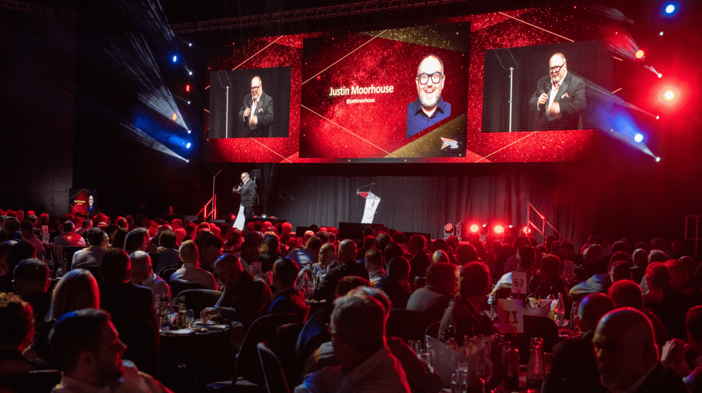A speaker stands on stage at a Live Expo awards event, addressing a large seated audience amid red and gold lights. Two screens behind him display his image and the name Justin Moorhouse.