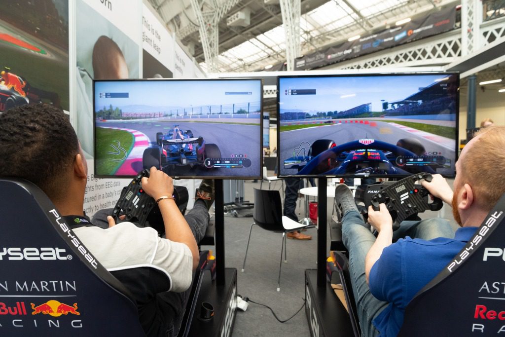 Two people sit in racing simulator seats with steering wheels, playing a car racing video game on large screens at an indoor experiential event. The screens showcase detailed race tracks and cars in action.