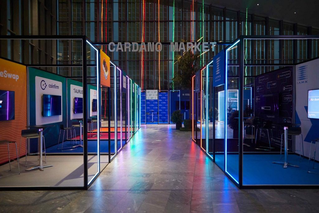 Colorful illuminated booths line both sides of a modern indoor exhibition space labeled “Cardano Market,” creating an immersive setting for experiential events. Digital screens and stools fill each booth, with vertical neon lights decorating the glass wall behind.