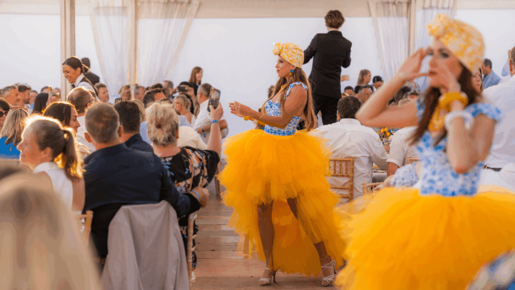 Two women in bright yellow tulle skirts and headwraps perform at a vibrant indoor sales incentive event, entertaining a seated crowd of guests at decorated tables in a festive atmosphere.