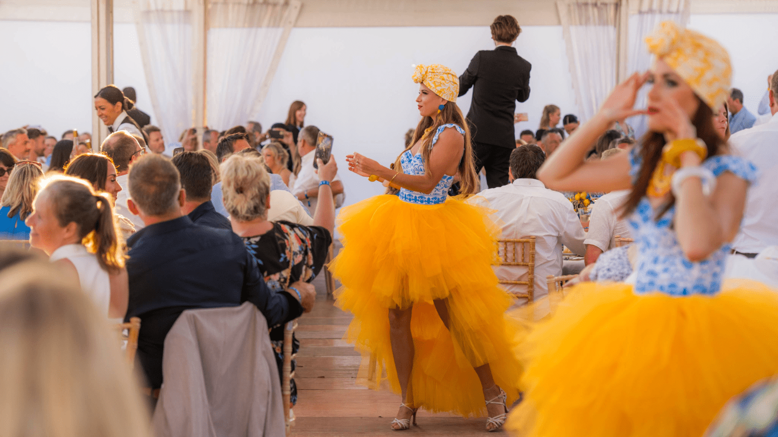 Two women in bright yellow tulle skirts and headwraps perform at a vibrant indoor sales incentive event, entertaining a seated crowd of guests at decorated tables in a festive atmosphere.