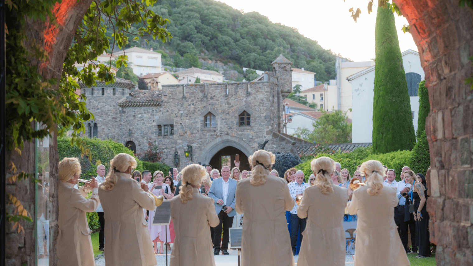 A group of musicians in white wigs and period costumes perform outdoors, celebrating a sales incentive, for an audience in front of a historic stone building and lush green hillside. The scene is framed by an arched entrance.