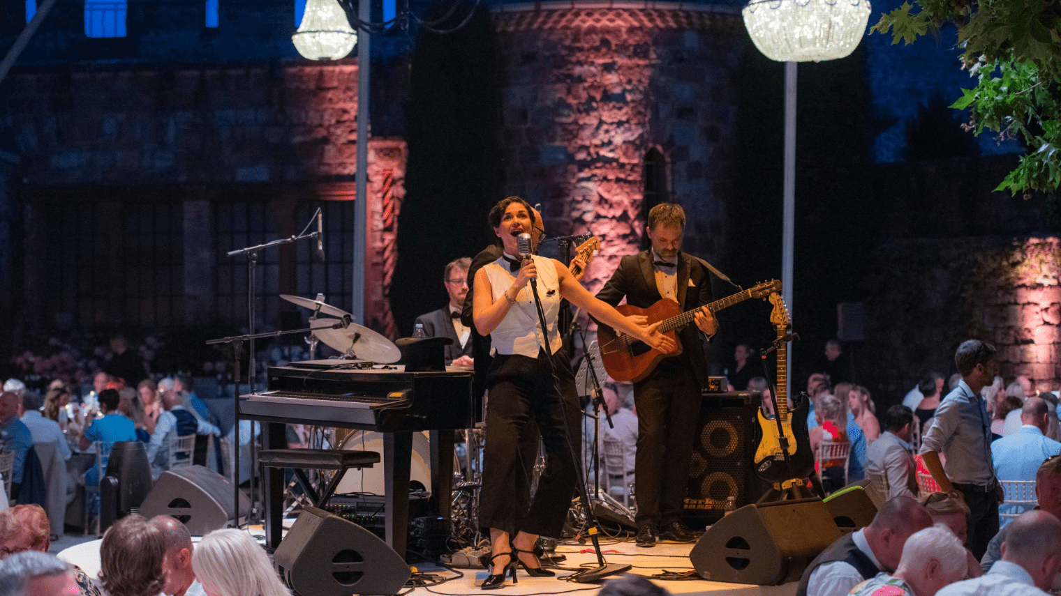 A woman sings into a microphone on stage with a live band, including a guitarist and drummer, performing at an elegant indoor sales incentive event with seated guests and chandeliers overhead.