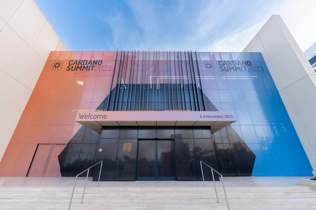 The entrance of a modern building with a glass facade, designed by a creative event agency, features the text Cardano Summit 2023 and event dates, with stairs leading to double doors under a pink Welcome sign.