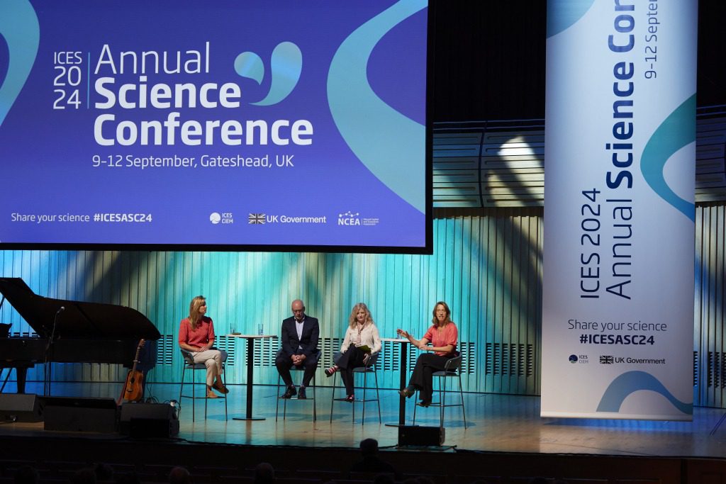 Four people sit on stage in front of a large screen at the ICES 2024 Annual Science Conference & exhibition in Gateshead, UK. The well-lit stage features banners displaying conference information and hashtags.