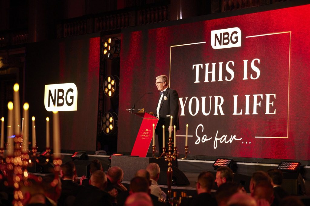 A man in a tuxedo speaks at a podium on stage during an event by a creative event agency. Behind him, a large red screen displays “NBG THIS IS YOUR LIFE So far...” Candles and audience members are visible in the foreground.