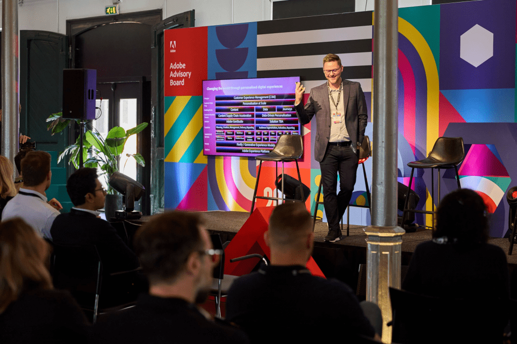 A man stands on stage, smiling and gesturing while presenting a slide to an audience at an Adobe Advisory Board event organized by a creative event agency. The stage features colorful geometric designs and empty chairs beside him.