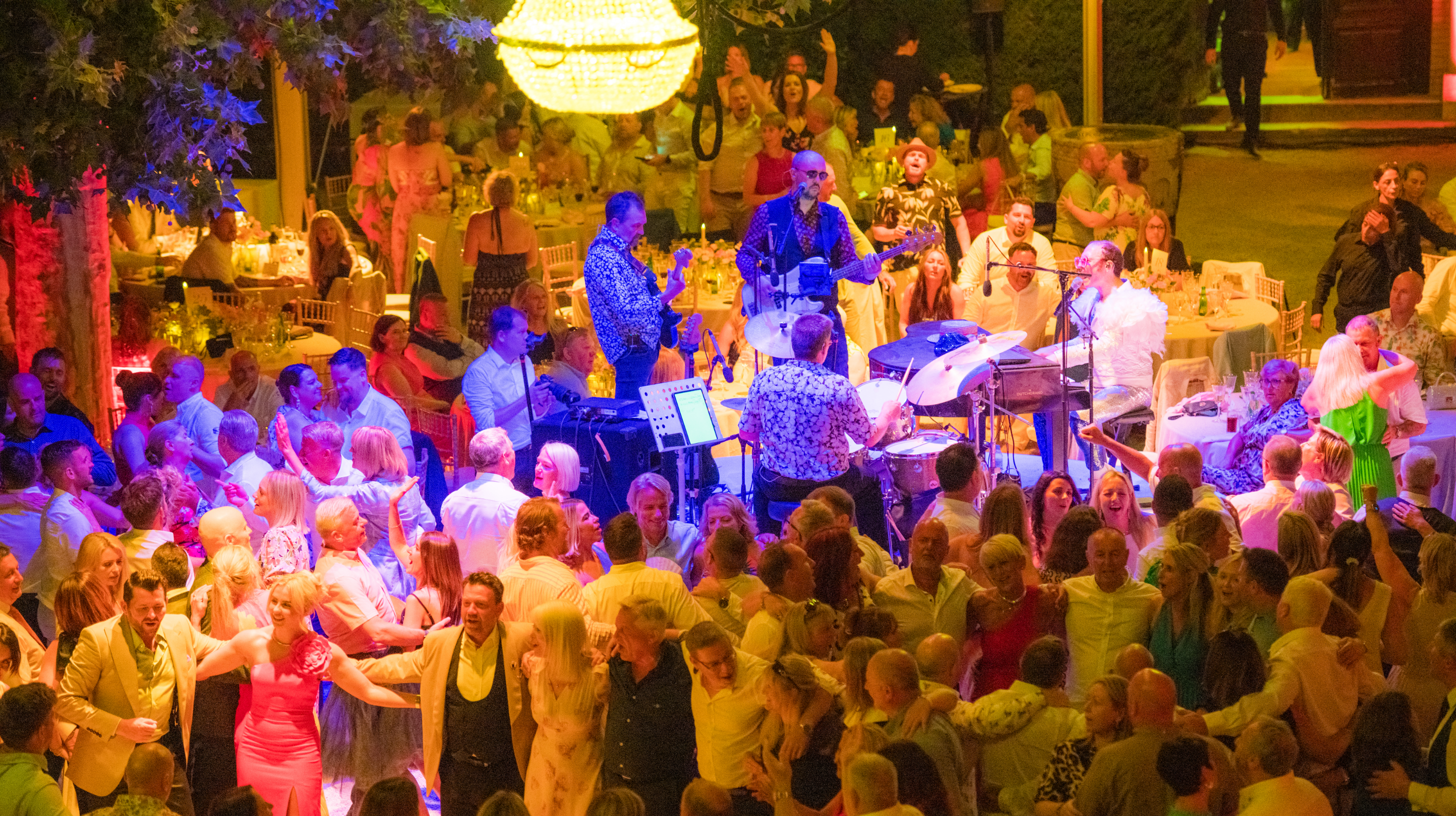 A lively crowd dances around a live band performing on stage at a brightly lit outdoor evening event, where guests in festive attire celebrate a successful sales incentive with tables buzzing with excitement.