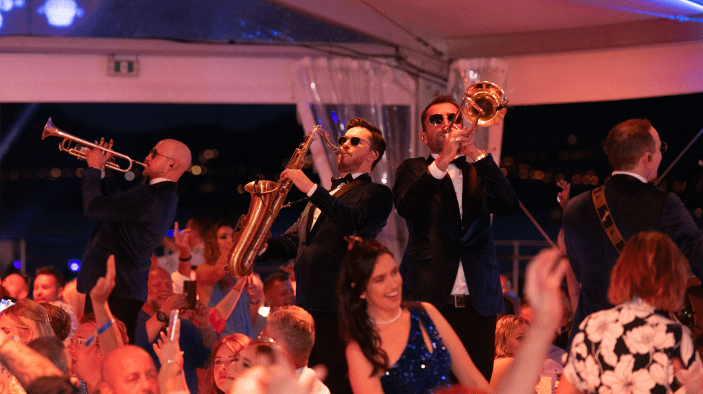 A lively band in suits plays trumpet, saxophone, and trombone on stage at a crowded sales incentive event, with an enthusiastic audience clapping and enjoying the music.