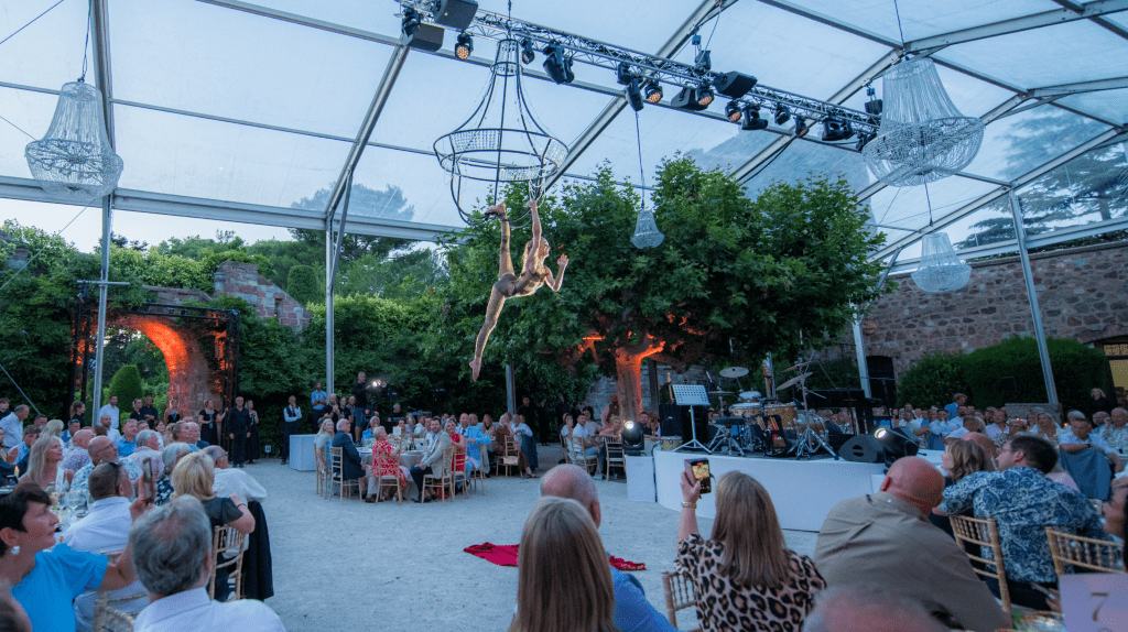 An acrobat performs on aerial silks suspended from a chandelier in a transparent tent, captivating an audience seated at tables for a unique sales incentive event. Trees and stone walls surround the spectacular venue.