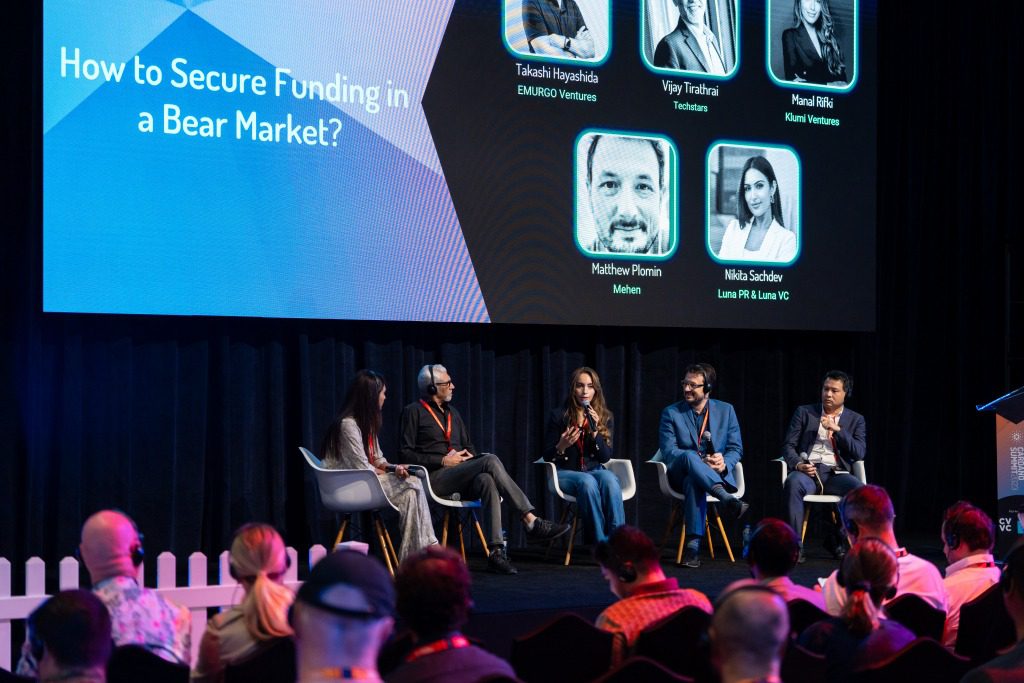 A panel discussion titled “How to Secure Funding in a Bear Market?” features five speakers on stage with a large screen showing their names and photos. This dynamic setup reflects the kind of programming and speaker formats shaping event trends 2025.