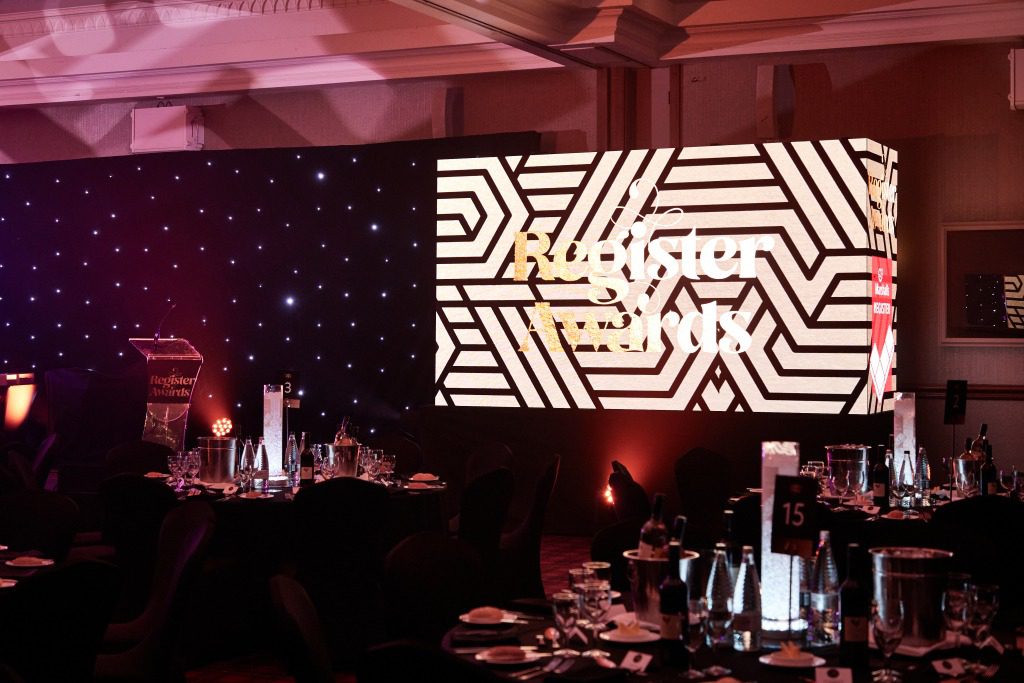A dimly lit banquet hall showcases thoughtful event design, with round tables adorned with bottles and glasses, and a large illuminated geometric-patterned screen displaying Register Awards.