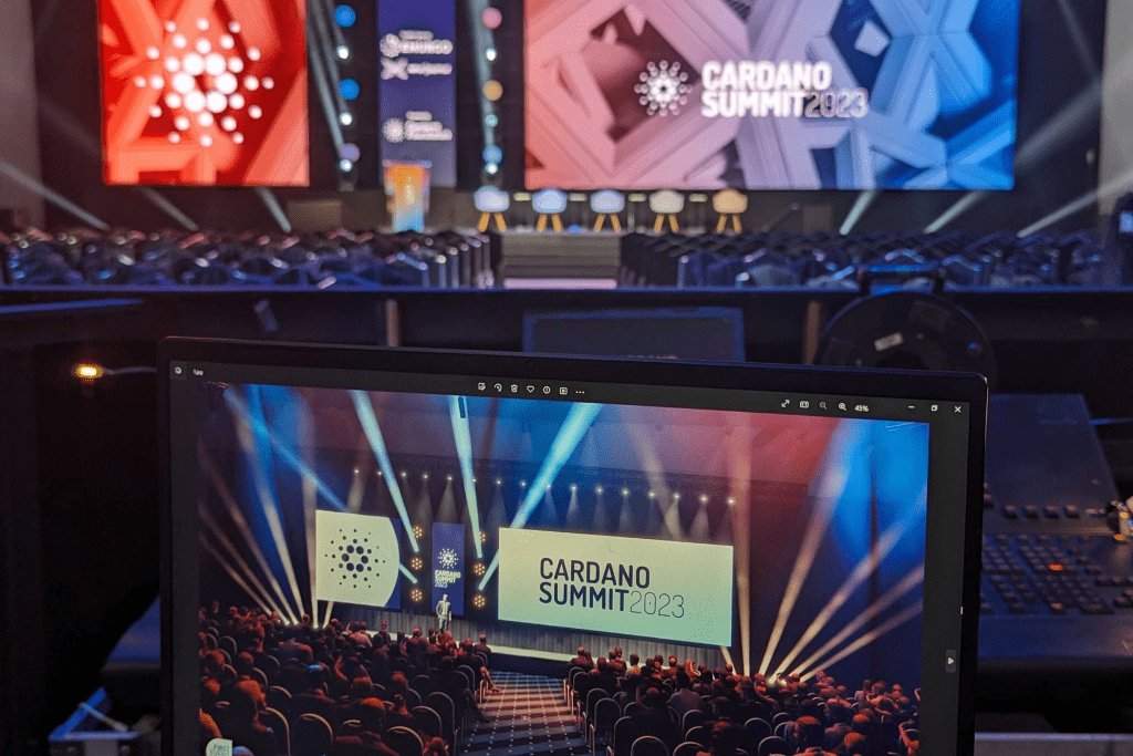 A photo of an event stage set for the Cardano Summit 2023, showcasing creative event design with colorful lights, empty chairs, large screens, and a laptop in the foreground displaying a similar scene.