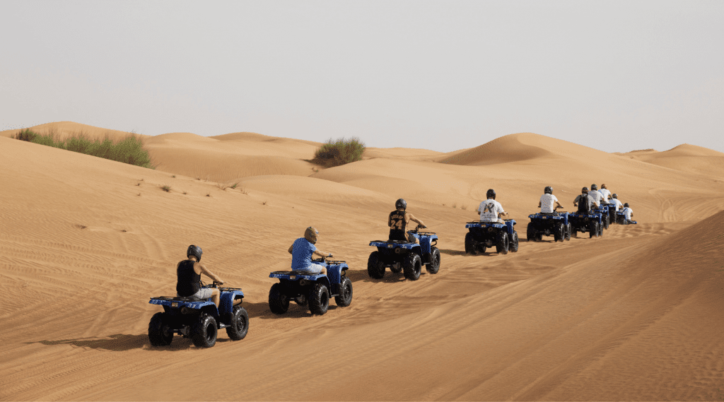 A group of people ride blue quad bikes in a line across sandy desert dunes under a clear sky, exploring sparse green bushes in the background—an exciting incentive event Dubai has to offer.