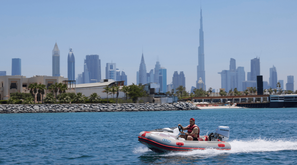 A man wearing a red cap and life vest rides a small inflatable motorboat on blue water during an incentive event Dubai, with a modern city skyline featuring skyscrapers in the background.
