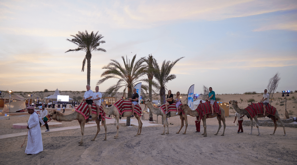 A group of people ride camels covered in red-striped blankets during an incentive event Dubai, led by a man in traditional white attire. Palm trees, desert sand, and glowing tents create a magical sunset backdrop.
