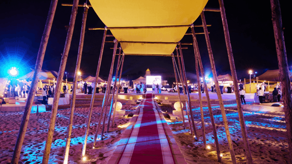 A nighttime outdoor incentive event Dubai on sand features a red carpet walkway under a canopy, flanked by poles and illuminated by blue and yellow lights, with guests mingling among tents in the background.
