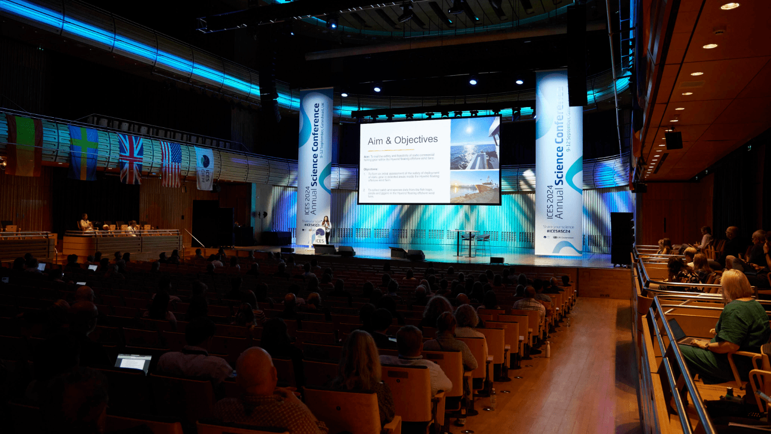 A speaker presents at a science conference in a large auditorium, with attendees seated, international flags on the left wall, and a large screen on stage displaying Aim & Objectives.