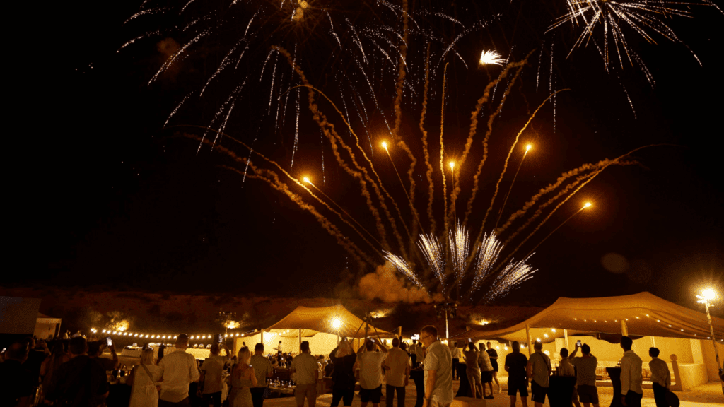 A crowd of people stands outside at night during an incentive event Dubai, watching colorful fireworks burst in the sky above brightly lit tents. The scene appears festive and celebratory, with warm lights illuminating the area.