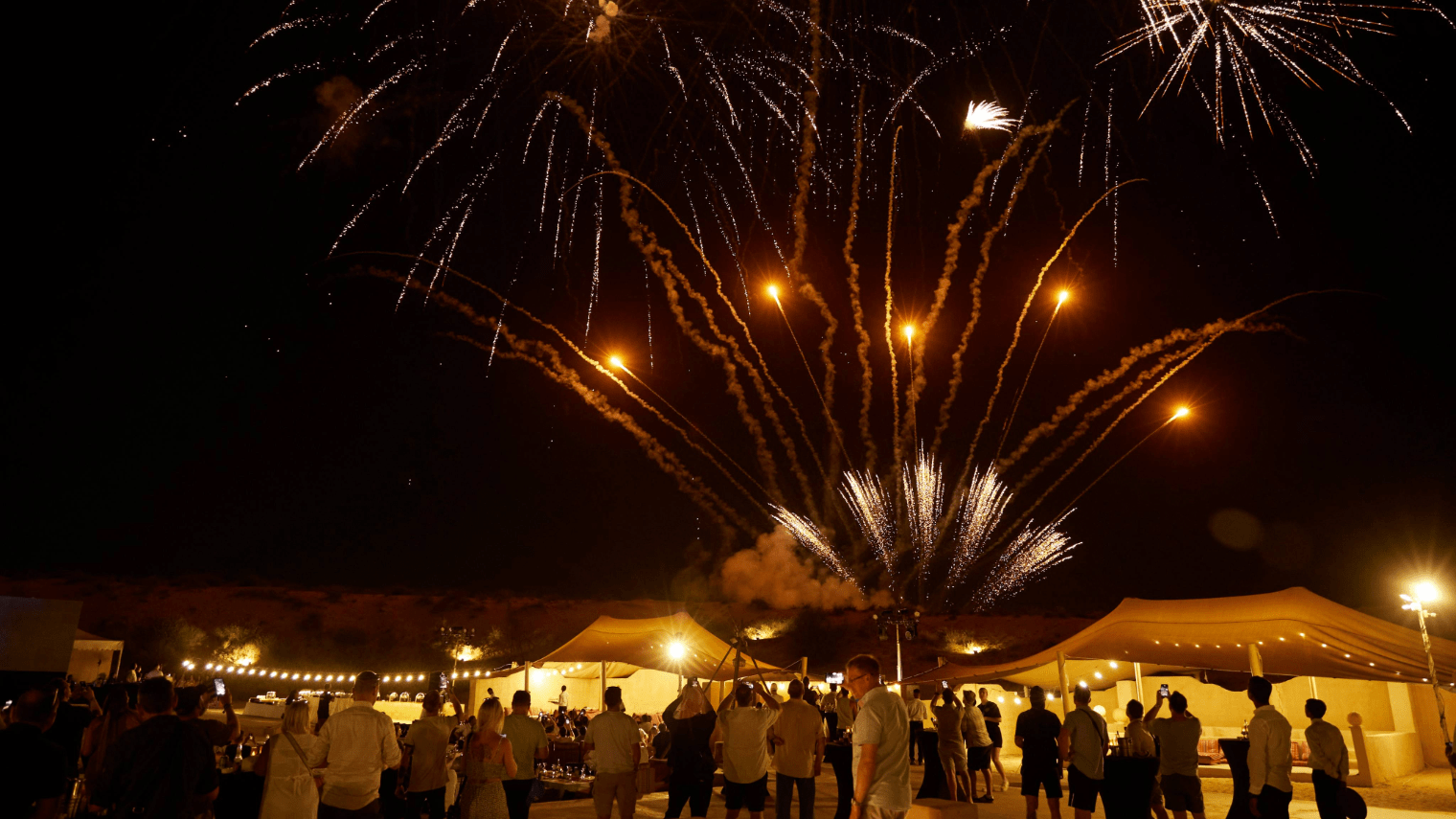 A crowd of people stands outside at night during an incentive event Dubai, watching colorful fireworks burst in the sky above brightly lit tents. The scene appears festive and celebratory, with warm lights illuminating the area.