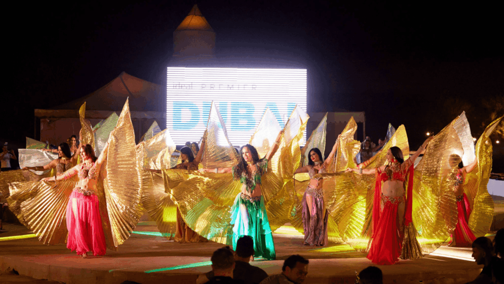 A group of women in colorful costumes perform a traditional dance with large, gold, fan-like wings on an outdoor stage at night during an incentive event in Dubai, with an illuminated sign and audience in the background.