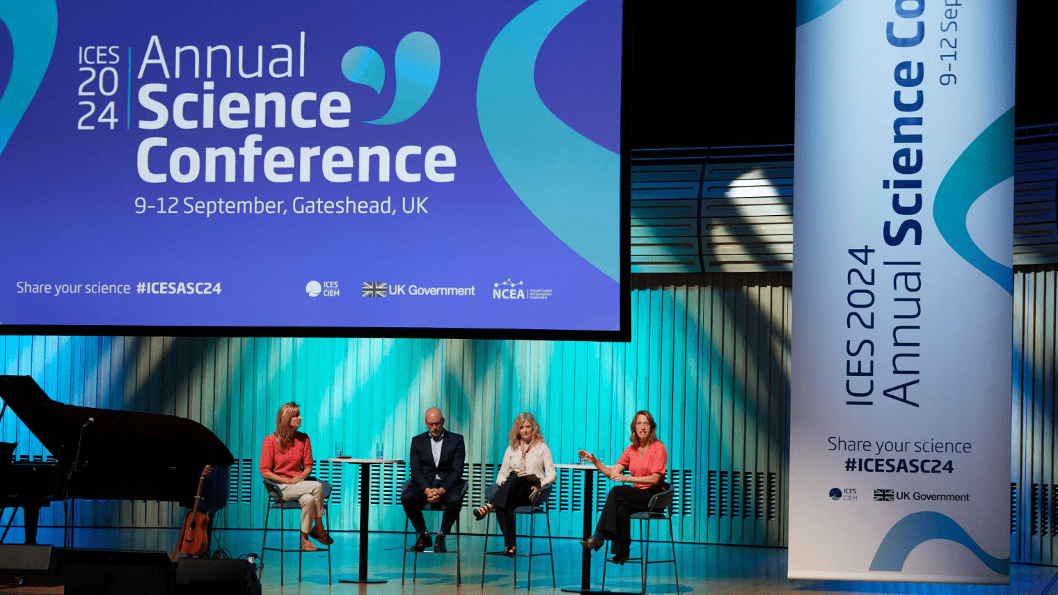 Four people sit on stage in front of a large sign for the ICES 2024 science conference in Gateshead, UK. A vertical banner and screen display event details as panelists speak to the audience.