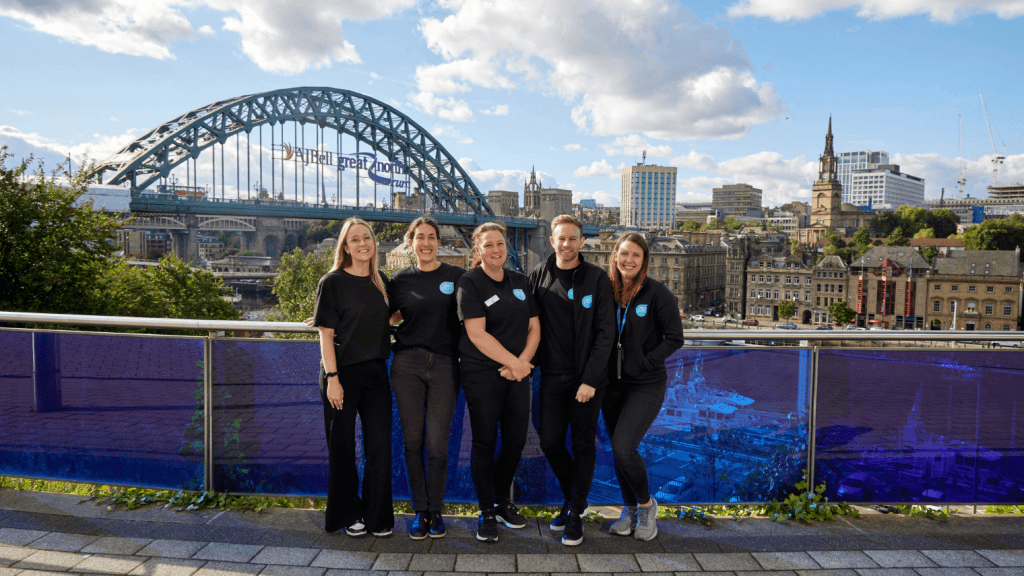 Five people stand smiling in front of a railing with the Tyne Bridge and Newcastle cityscape in the background on a sunny day, enjoying a break during the science conference.