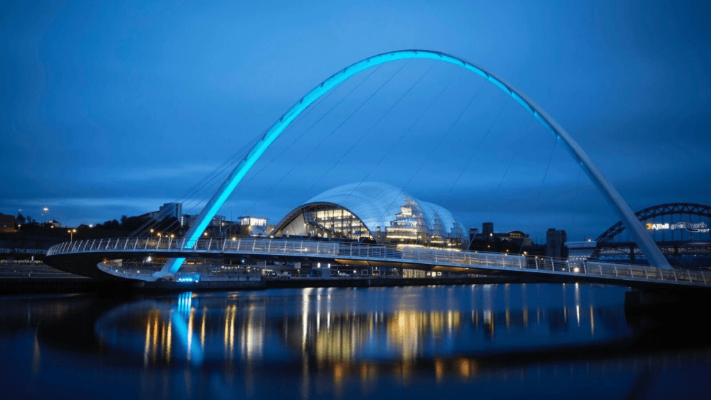 A modern arched pedestrian and cyclist bridge with blue lights spans a calm river at dusk, leading toward city buildings and an illuminated glass-domed structure, evoking the energy of a bustling science conference in the background.