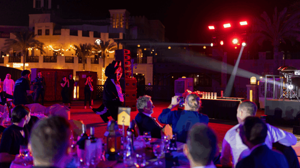 An outdoor blockchain event at night with people seated at tables, some snapping photos of a performer on stage. Bright stage lights and festive string lights create a lively vibe, with bottles and glasses on the tables.