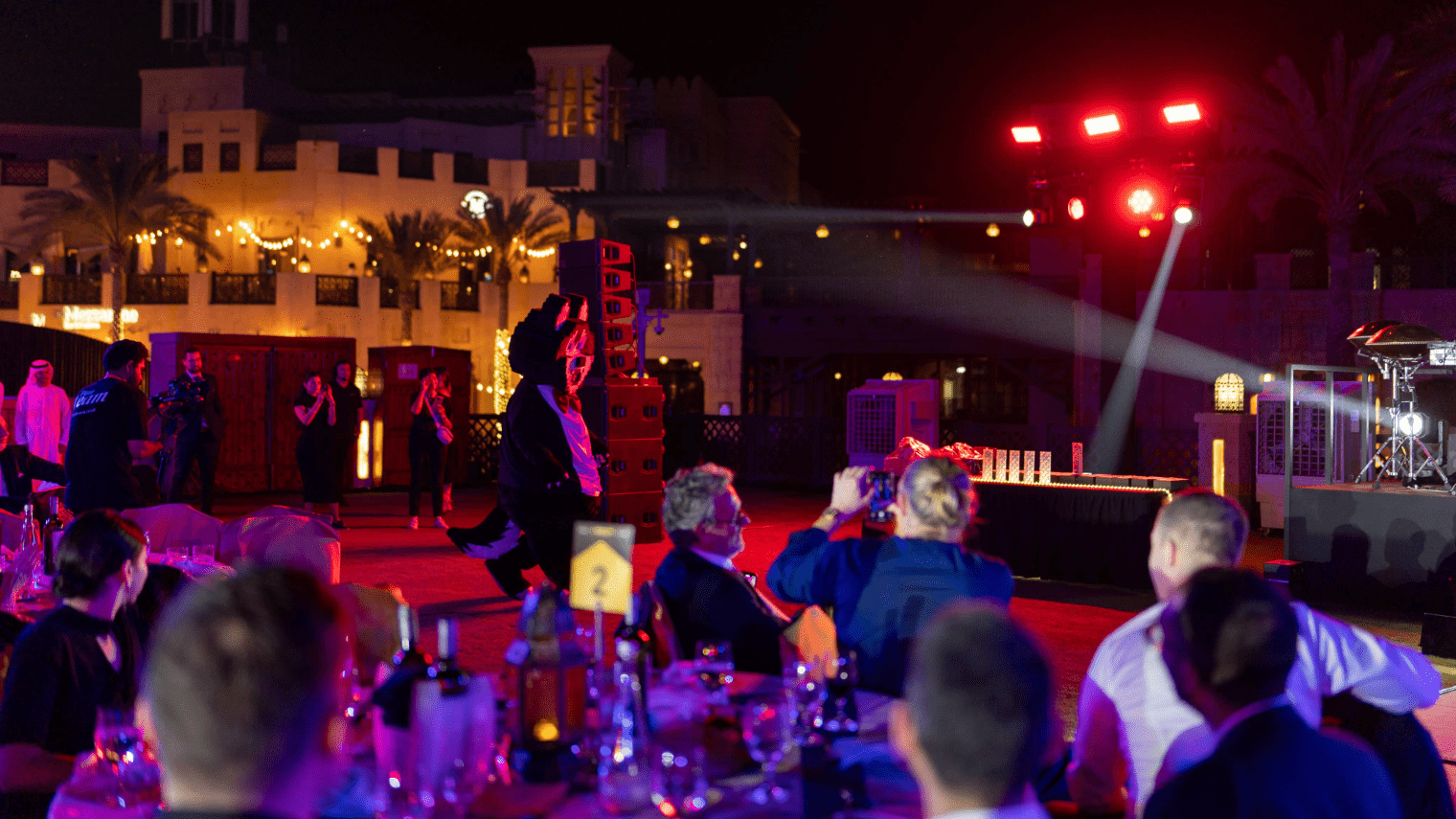 An outdoor blockchain event at night with people seated at tables, some snapping photos of a performer on stage. Bright stage lights and festive string lights create a lively vibe, with bottles and glasses on the tables.