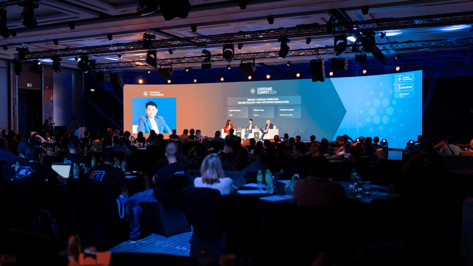 A blockchain event conference panel features speakers on stage, a large screen displaying a speaker’s image, and audience members seated at tables in a dimly lit room bathed in blue lighting.