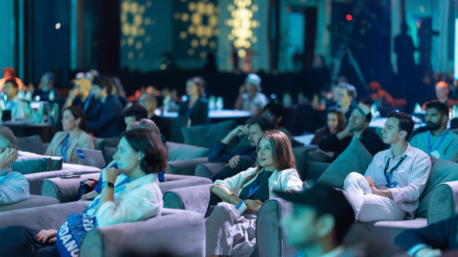 Audience members sit in cushioned chairs, attentively watching a blockchain event presentation in a dimly lit conference room with blue lighting, focused and engaged—some taking notes, others listening intently.
