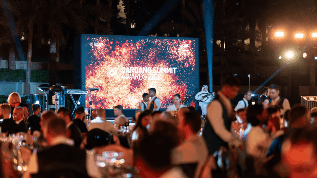 Guests seated at tables during an outdoor evening blockchain event, with servers walking around. A large screen in the background displays “Cardano Summit Awards 2023” with a sparkling graphic.