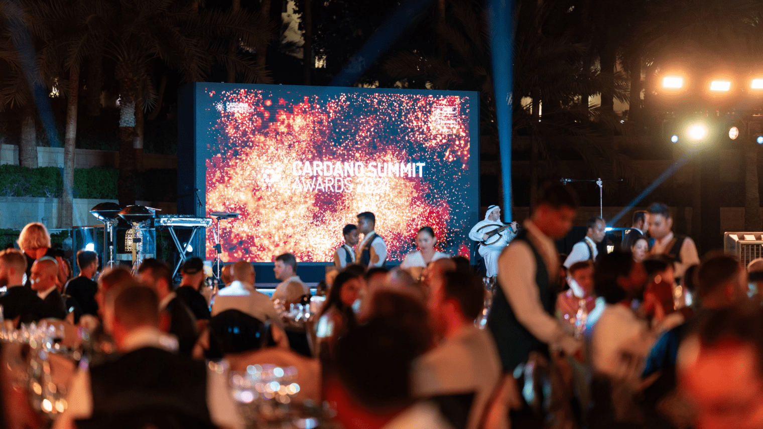 Guests seated at tables during an outdoor evening blockchain event, with servers walking around. A large screen in the background displays “Cardano Summit Awards 2023” with a sparkling graphic.