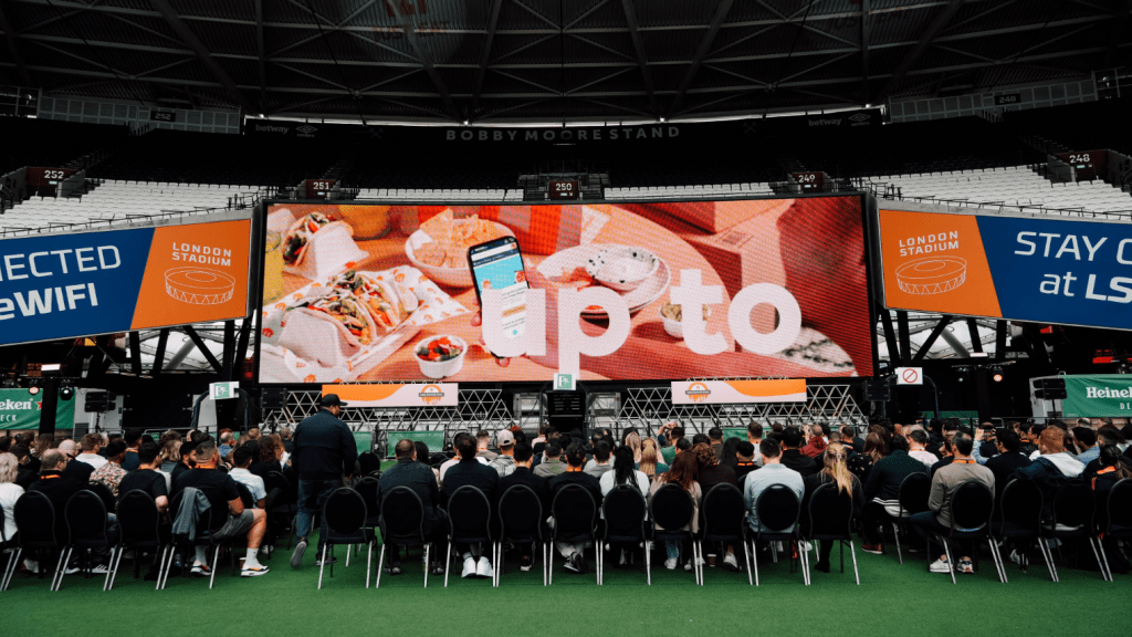 A crowd sits facing a large screen displaying food and the text up to inside a stadium, likely during the London Conference. Digital side screens read STAY CONNECTED AT LS WIFI and LONDON STADIUM. The stadium roof and seating are visible.