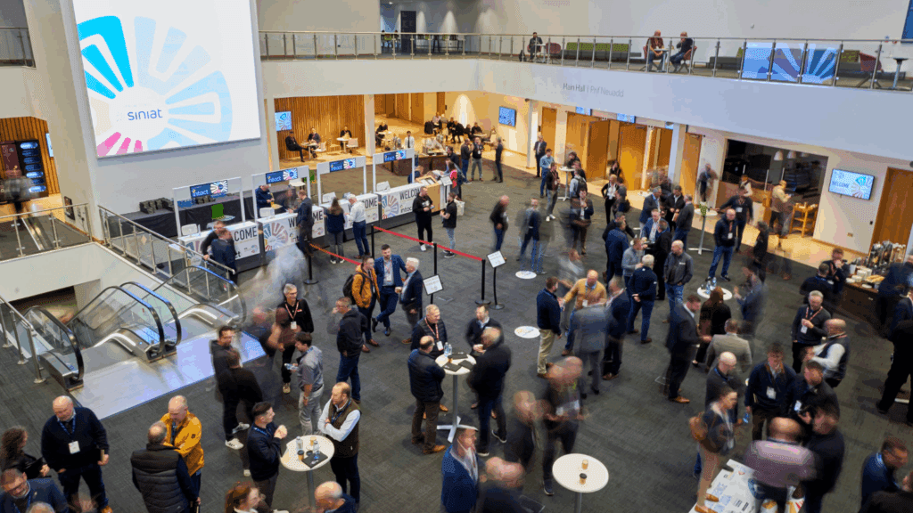A busy conference & exhibition hall with people mingling, talking, and networking. Several booths and tables fill the space, while a large screen with the Sinat logo is displayed on the wall. Escalators are visible to the left.