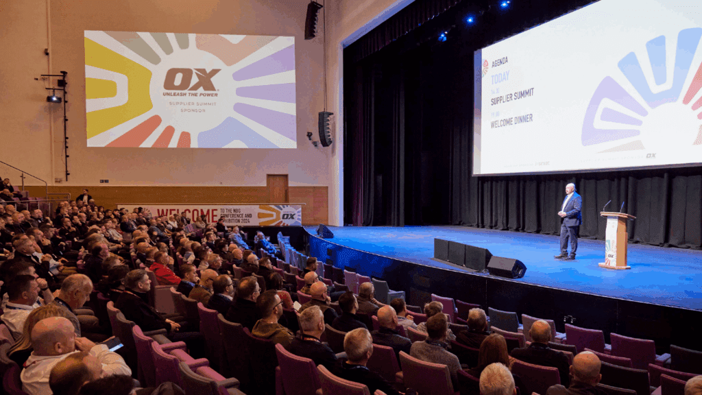 A speaker stands on stage at a conference & exhibition, addressing a large audience in an auditorium. Colorful graphics and text, including “OX,” appear on a screen behind the speaker and on the wall to the left. The audience sits attentively.