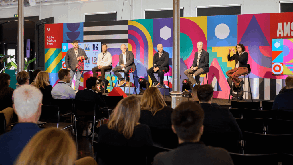 A panel of six speakers, including one using sign language, sits on stools on a colorful stage with geometric designs, sharing insights to enhance the experience for digital creatives at an Adobe Advisory Board event.
