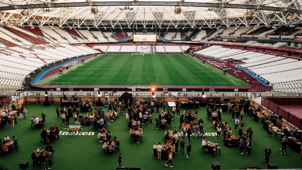 A large group of people gather on a green floor area for a London Conference event inside a mostly empty stadium with white seats and a well-maintained soccer field in the background.