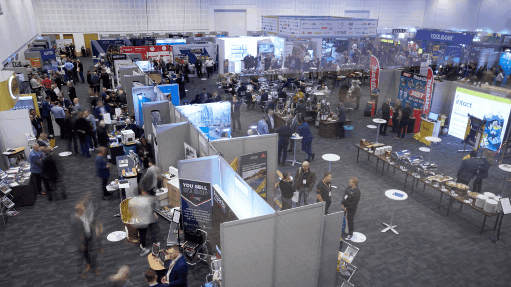 A busy conference & exhibition with many booths, displays, and people networking or exploring products in a large, brightly lit hall. Some attendees are seated at tables, while others walk between exhibits.