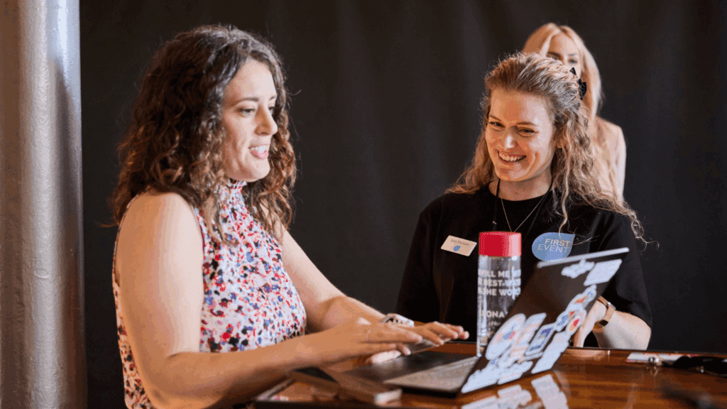 Two women are smiling and talking at a table with an open laptop covered in stickers. One types while the other watches, coffee tumbler in hand—an inspiring scene highlighting the collaborative experience for digital creatives.