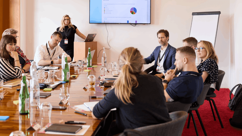 A group of people sits around a conference table in a meeting room, listening to a woman presenting near a screen displaying a chart—an engaging experience for digital creatives. Laptops, notebooks, and water bottles are on the table.