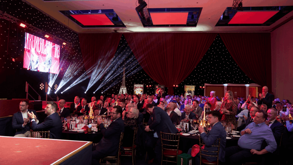 A large group of people in formal attire sit at round tables during an indoor gala, conference & exhibition or awards ceremony, with stage lights, a red curtain backdrop, and a presenter visible on stage.