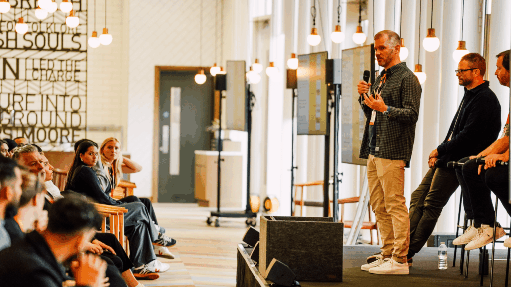 A man stands and speaks into a microphone on stage at the London Conference, addressing a seated audience. Two other men sit beside him in a modern setting with large windows and hanging lights as the audience listens attentively.