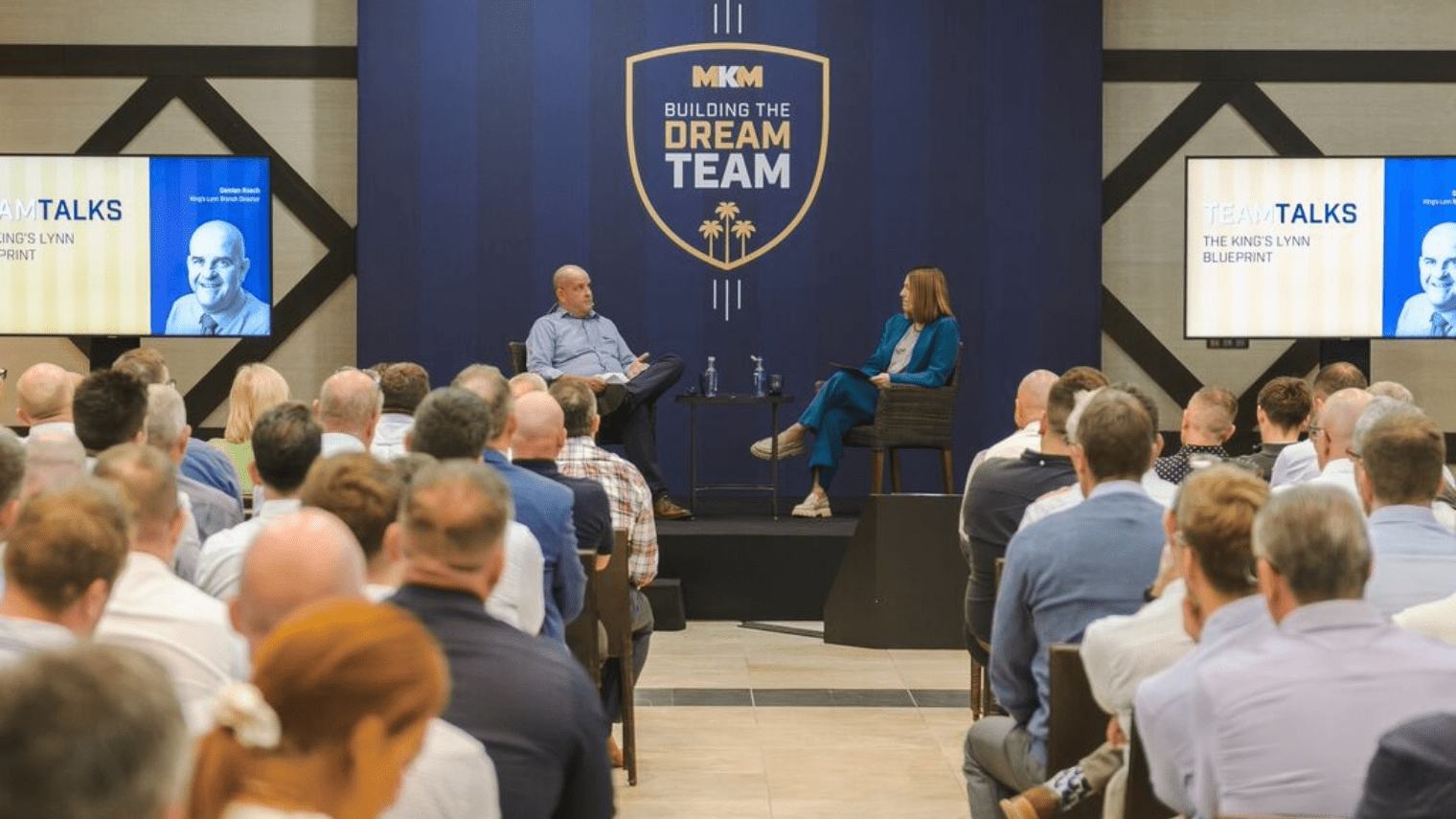 A man and a woman sit on stage having a discussion in front of an audience at a strategic conference titled “BUILDING THE DREAM TEAM.” Two screens display “TEAM TALKS” and a headshot of a speaker.