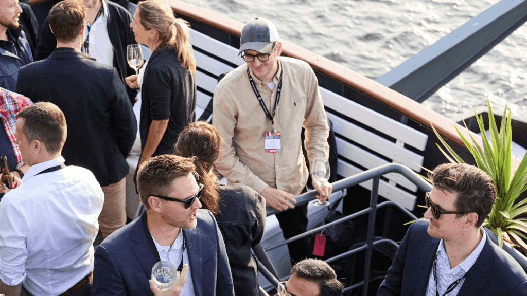 A group of people socialize on a boat deck by the water—some holding drinks and wearing lanyards, with a mix of casual and business attire—enjoying an inspiring experience for digital creatives.